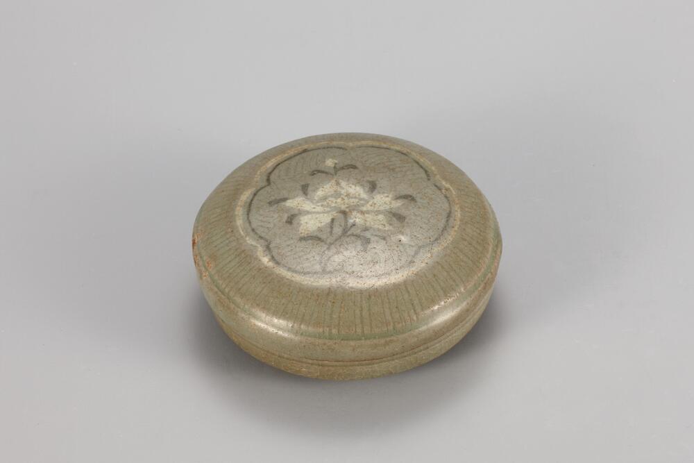 <p>This is a celadon lidded bowl with the top of the lid decorated with a peony spray design inside a hexafoil outline inlaid with black and white slips. The foot was made by removing clay from the underside of the base and retains traces of quartzite spurs in three places. The glaze was slightly darkened on the upper part with faint gloss. The glaze is poorly fused on the base of the body, leaving practically no sheen. The piece is of high value, however, for the glimpse it offers of Goryeo&rsquo;s refined yet splendid inlaid celadon ware, thanks to its decorative design that depicts a peony in full bloom, inlaid with white clay.<br />
[<em>Korean Collection, University of Michigan Museum of Art </em>(2014) p.118]<br />
&nbsp;</p>

