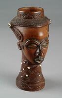 This richly detailed, carved wooden cup has been sculpted into the form of a human head. The head&rsquo;s bell-shaped coiffure and facial details are typical of Kuba masks and figurines. The face features almond-shaped eyes, a protruding mouth and nose, disproportionately small ears, and eyebrows and temples that have been engilded with tiny copper staples. The neck and the coiffure bear elaborate diamond-shaped and diagonal-lined patterns which have been further embellished by cowrie shells, embedded in resin.