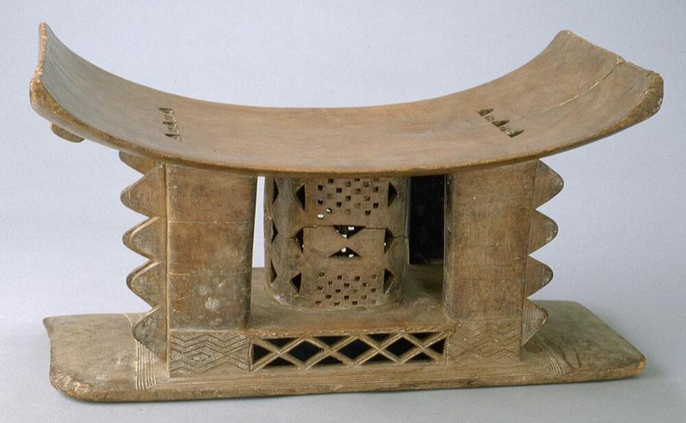 Wooden stool with a rectangular base and a central column supporting a rectangular seat that curves upward at each end. The central column is decorated with open-work designs of triangles and a checkerboard pattern. Surrounding the column are four rectangular posts decorated with triangular projections along the outer edges. 