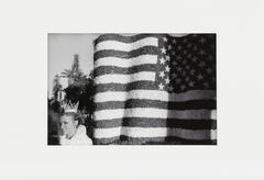 A black and white image of the reverse of an American flag, curled and appearing textured and solid. To the left bottom, a woman with a crown and a white jacket looks towards the viewer.