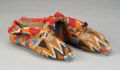 Shoe with a leather sole attached to a cloth lining with metal tacks. The outside of the shoe is covered in multicolored beadwork; the toe has a pattern of zig-zags in orange, black, and white while the sides and upper flaps have an interlacing pattern in cream, red, blue, and orange. The top of the shoe is bound with red cloth along the edge.