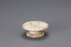 A small glazed porcelain piece resembling a cake stand. The top is detailed with a carved pattern that would imprint the rice cake being molded on it.<br />
<br />
Rice cake stamps are used to impress designs upon rice cakes. They are generally made of wood or ceramic. Ceramic rice cake stamps normally come in the form of round stamps and consist of a patterned surface and a handle. Patterns, carved or raised, on the stamp vary from geometric lines to auspicious designs that wish for prosperity and longevity. Their small size makes them highly portable, while their simple yet contemporary designs have mad them popular among collectors. The University of Michigan Museum of Art collection includes nine white porcelain rice cake stamps. Some are gifts from Mr. and Mrs. Hasenkamp, and others are gifts from Ok Ja Chang and the Chang family.<br />
<br />
Glaze has been removed from the foot rim, on which fine sand spur marks remain.<br />
<br />
[Korean Collection, University of Michigan Museum of Art (2014) 