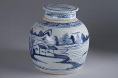 Blue-and-white covered jar with landscape design of a river, trees, mountain, houses and it has a lid with blue an white designs.