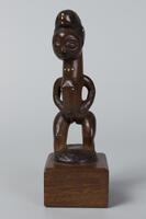 Standing nude female figure with bent knees. The carved hair has a criss-cross pattern and there are bead inlays on the figure&#39;s hair, neck, and breasts. The figure&#39;s hands are at its hips and the bottom of the piece is an oval connecting to the figure&#39;s ankles in place of feet.