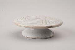 A small glazed porcelain piece resembling a cake stand. The base is wide and short and sharply tapered inward where it meets the top. The top is detailed with a carved pattern that would imprint the rice cake being molded on it.<br />
<br />
Rice cake stamps are used to impress designs upon rice cakes. They are generally made of wood or ceramic. Ceramic rice cake stamps normally come in the form of round stamps and consist of a patterned surface and a handle. Patterns, carved or raised, on the stamp vary from geometric lines to auspicious designs that wish for prosperity and longevity. Their small size makes them highly portable, while their simple yet contemporary designs have mad them popular among collectors. The University of Michigan Museum of Art collection includes nine white porcelain rice cake stamps. Some are gifts from Mr. and Mrs. Hasenkamp, and others are gifts from Ok Ja Chang and the Chang family.<br />
<br />
&nbsp;This mold is entirely glazed, but it is has been removed from the foot rim.