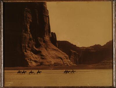 A team of seven people on horses and a dog cross the desert. They appear small compared to the vast rocky landforms behind them.