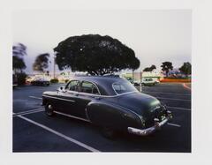 Image of a black sedan in a parking lot. Behind the car are trees, three more cars, and a green and white bus. 
