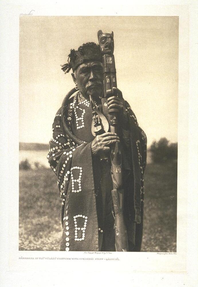This is a portrait of an aging man in an outdoor landscape. He holds a large, carved wooden staff, which portrays an eagle at the top, and other faces and forms throughout the design. He wears an embroidered cloth draped over his body, a hat, and jewelry.