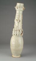A tall porcelain vase with an ovoid lower body, a tall narrow neck, and dish shaped mouth on a footring. The neck has several applied figures of molded immortals and sculpted cosmological creatures, and four loop lugs. It is covered in a white glaze with a bluish green tinge. The lid is missing.