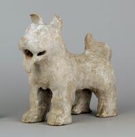 A red earthenware dog sculpture.  The dog is standing in attention, with a stocky body on four muscular legs, and long-fur tail flowing up at the hindquarters.  Its head is atop a thick neck, and it has a smiling mouth with protruding tongue, deep-set large eyes and forward pointed bent ears.  Its pupils are detailed in black. There are traces of black and white mineral pigment. 