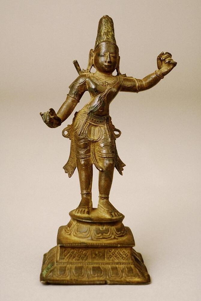 The two-handed figure stands in relaxed posture on a base consisting of square and round forms.  At the bottom of each shape stylized lotus petals are incised.  His right hand is extended outwards and would have been holding an arrow and he left arm is extended up to his die to hold a bow.  He wears much of jewelry including anklets bracelets, armlets, necklace and shoulder loops, belts with pendant elements and a sacred thread reaching just below his waist.  He wears a lower garment with incised lines delineating the folds and sections that flare out to either side.  He wears an elaborate crown.  One element extends over his right shoulder, which may either be part of his quiver of arrows or a broken section of a halo. He wears a conical headdress incised with a worn pattern.  His face is sharply stylized with a long straight nose and wide, open eyes.<br />