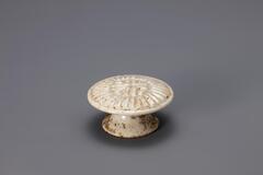 A small glazed porcelain piece resembling a cake stand. The base is wide at the bottom and slopes inward towards the base of the top. It also has a hole cut out of it on one side. The top is detailed with a carved pattern that would imprint the rice cake being molded on it.<br />
<br />
Rice cake stamps are used to impress designs upon rice cakes. They are generally made of wood or ceramic. Ceramic rice cake stamps normally come in the form of round stamps and consist of a patterned surface and a handle. Patterns, carved or raised, on the stamp vary from geometric lines to auspicious designs that wish for prosperity and longevity. Their small size makes them highly portable, while their simple yet contemporary designs have mad them popular among collectors. The University of Michigan Museum of Art collection includes nine white porcelain rice cake stamps. Some are gifts from Mr. and Mrs. Hasenkamp, and others are gifts from Ok Ja Chang and the Chang family.<br />
<br />
The clay body is exposed at the foo