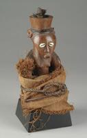 This woodcarved figure depicts a female with a large bowl-shaped vessel upon the crown of her head, intended to be a container for symbolic, medicinal and spirit “activating” ingredients. Representative of the Kasongo style (regarded as the “classical” or “pure” Kusu stylistic form), this <em>kakudji</em> features an inverted, triangular-shaped head, a wide, convex forehead, high ears, cowrie shell eyes, a triangular nose, an oval mouth with protruding lips, a pointed chin, and conical breasts. The figure possesses a prominent belly indicating pregnancy, and representing the themes of maternity, fertility, and the continuation of the lineage. A large piece of textured cloth tied in place with rope encircles the female’s lower arms and lower body.