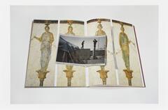 Image of a book lying open to a page featuring frescos depicting figures on pedestals. A newsclipping featuring a photo of a person standing on a pole lies on the book across the open page.  
