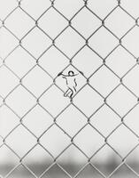A black and white photograph of a linked fence. Drawn into the center link is a figure with hands around the upper fencing.