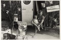 A black and white image of a man sitting and holding a child in front of a trailer. To the left is a sign that reads 'DIVORCE SALE! EVERYTHING GOES!'