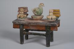 This is an earthenware miniature table with black legs, glazed in brown and amber. It has five objects placed on the table in two rows - three in back and two in front. In the back row is a central bowl holding a green duck, flanked by two other bowls holding what looks like logs of firewood and split bamboo. In the front row are two bowls stacked high with round fruit. 