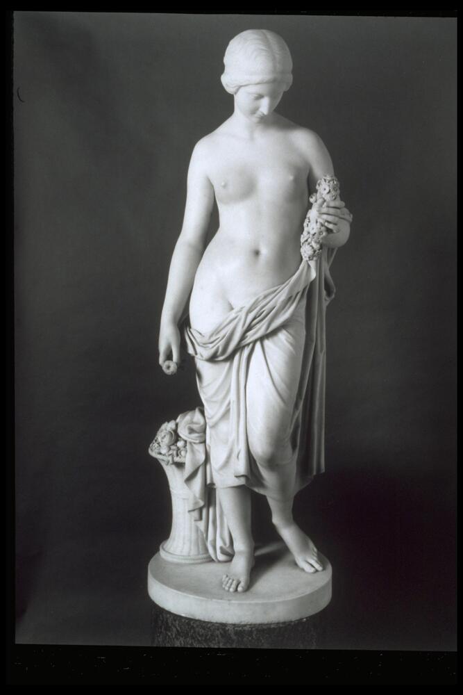 White marble sculpture of female figure, partially nude with a cloth draped loosely around her waist and over her left forearm. She holds a cluster of flowers in her left hand, and a single bloom in her right; a basket of flowers located on base to left and slightly behind figure.