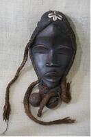 Oval black face mask articulated, protruding angular mouth, nose, and eyes with eye slits. The figure has cowrie shells at the top of the forehead with braided raffia hair on either side of the head and bells beneath the chin.&nbsp;