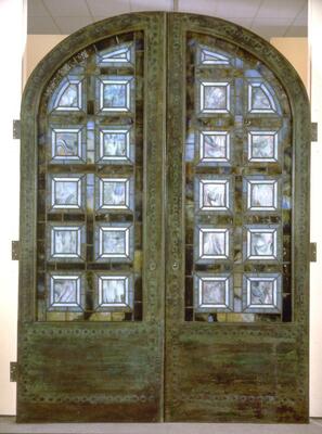 One of a pair of doors that formed an arched entryway. In the upper two thirds of the door are opalescent square glass &quot;coffers&quot; in an arched composition that corresponds to the silhouette of the doors. The interior-facing side of the doors include curvilinear lead caming, inset with medium-sized beach stones, that frame the glass &quot;coffers&quot;. The exterior-facing side of the doors has the &quot;coffers&quot; framed by patinated copper sheeting.