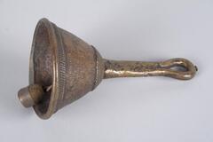 Hand-held bell with a short handle ending in a loop. The dome-shaped bell has a pattern of incised lines around the top edge and a grid-like pattern around the lip. Inside the bell hangs a small clapper. 