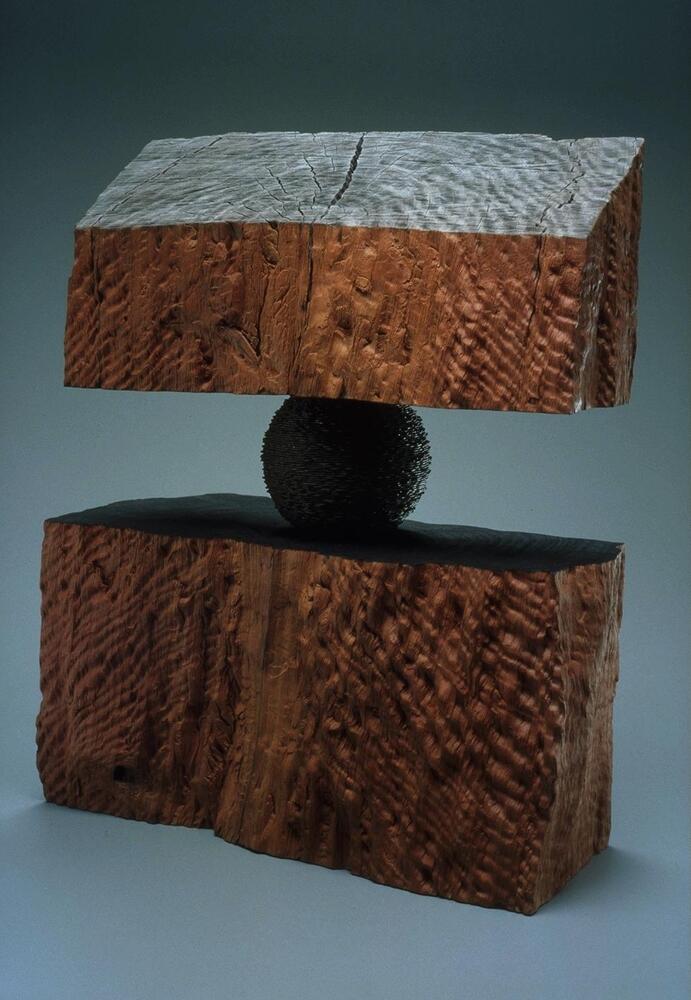 A sphere, wrapped in rusted wire, is sandwiched between two hefty, rough hewn, chunks of wood.<br />
freestanding multimedia wood sculpture, with rectangular wood base and top sandwiching a small wire-covered sphere at center