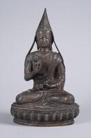 A very finely hollow cast bronze portrait sculpture of a seated figure, with the lotus dais and pointed monk's cap cast in one piece with the figure.<br />The monk is shown seated in the padmasana (lotus) pose, with each foot resting sole-upward on the opposite knee. In his right hand, he holds a vajra (a double-pronged scepter) and simultaneously makes the vitarka gesture for teaching. His left hand, resting on his lap, holds a bell. His costume consists of a dhoti, which is knotted high on his torso; a short-sleeved shirt, crossed over his chest and decorated with incised scroll patterns, with a fret design at the border; and an overrobe that wraps around his left shoulder and is draped over his right shoulder.  His face has a broad forehead, incised eyebrows in a high arch; downcast eyes, with leaf-shaped upper eyelids; a broad, flat nose; a sweet smile and full lips; and a narrow chin. His tall, pointed monk's cap, which completely hides his hair, has flaps that spread to reach his upper arms.