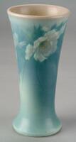Vessel is a tall vase with a flaring lip and a slight flaring at the base.  The overall glaze is a blue matte "vellum" glaze with underglaze decoration depicting a band of white peonies below the lip.  The lip itself modulates to a rosy pink color.