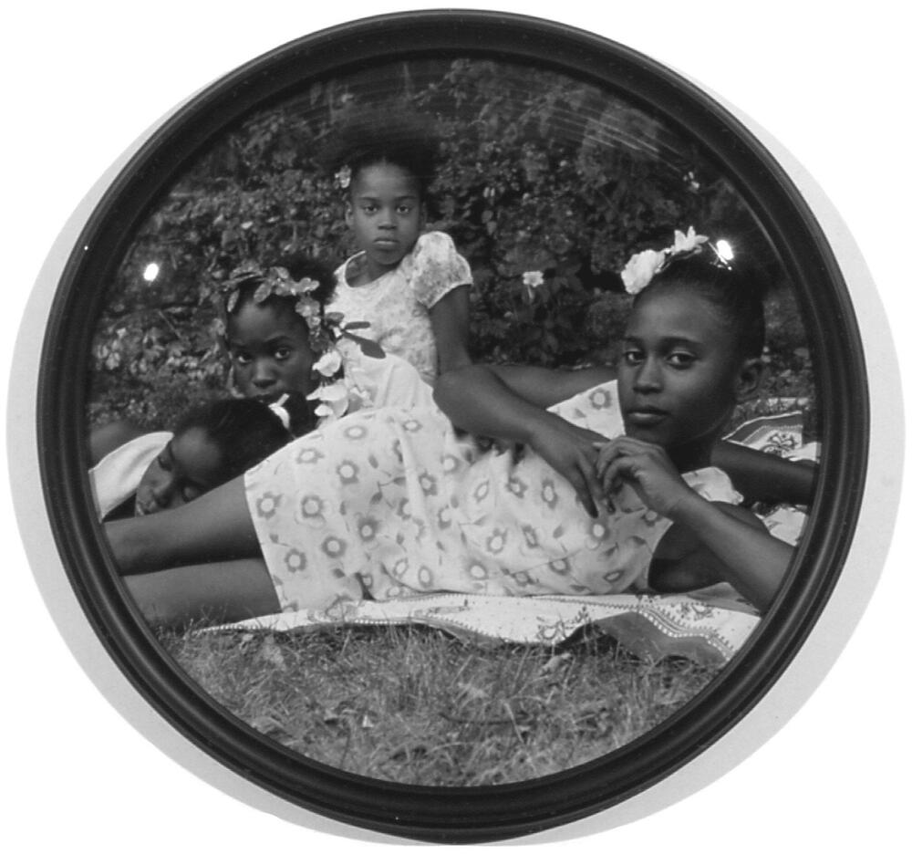 This image is a circular format black and white photograph depicting four young African American girls wearing floral dresses lounging on a blanket in the grass. Three girls sit or prop themselves up and look at the camera, the fourth girl lays down, with her eyes closed. The glass in the frame has a domed surface, creating a fish-eye effect.
