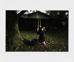 An image of a child dressed in black clothing, sitting on a tire swing with a yellow chain and turning to look at the viewer. In the dark background, a house is visible and leaves are scattered on the ground.