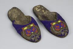 Open-back shoes with leather soles attached to a cloth-lining with metal tacks. The beadwork on the upper part of the shoe is blue with a British style crown rendered in gold, silver, red, blue, yellow, and green beads. 