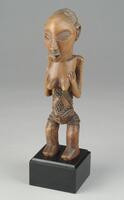 This standing female <em>nkisi mihasi</em>, or “benevolent” power figure, has been carved in light wood and exhibits many of the typical traits associated with the Luba Shankadi style, and more specifically, with the Mwanza center of production. These features include its disproportionately large, ovoid face, half open coffeebean-like eyes, wide mouth with full lips, triangular nose, high, convex forehead, cascading coiffure, protruding umbilicus, and, diamond-shaped tattoos carved in relief on the belly and in horizontal lines on the lower back and upper thighs. The figure stands with slightly flexed knees and with its arms bent at the elbows such that its palms rest upon the breasts, a pose commonly seen throughout Luba figural depictions of women.