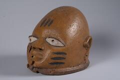Carved wood mask in the form of a human head. The head is bald, there are three black horizontal marks on each cheek, and three black vertical marks on the forehead. The pupils are pierced and the bottom of the mask has a raised lip. The inside top of the mask is filled with sand. 