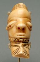 This small Pende ivory pendant (<em>ikhoko</em>) depicts a delicately carved male head. Stylistically, <em>ikhoko</em> bear many of the facial characteristics of anthropomorphic Pende <em>mbuya</em> masks, after which they have been modeled. In this particular example, an ovoid head rests atop a columnar neck and features a tall, striated coiffure.  It has a large, bulbous forehead, a V-shaped brow, heavily lidded and downcast eyes, an up-turned nose, a wide oval mouth, and a slightly pointed chin. Fine patterns and lined motifs decorate the neck. The pendant also possesses a pale yellow patina.