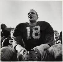 Image of a man sitting on a bench. He wears a jersey with the number &#39;18&#39; on it and holds a helmet in his lap.