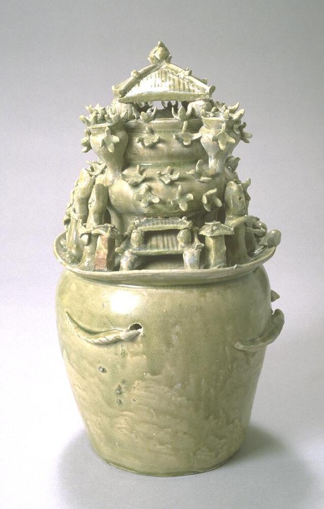 A large stoneware jar with a slightly tapered body, wide shoulders and a wide flaring mouth.  There are smaller jars stacked on top, getting progressively smaller, topped with a hipped roof mimicking contemporary architectural features and additional architectural features such as towers around the mouth and a plethora of assorted figures, birds, and cosmological animals applied around the top half of the vessel.  It is covered in a grey-green celadon glaze. 