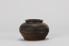 A gray-brown jar with a short, splayed neck. High-fired stoneware jar from the Goryeo or Josen period. The wave design on the side is set between two horizontal ridges.<br />
<br />
This is a gray-brown, high-fired stoneware jar. Its short neck splays sharply and is connected to a mouth that spreads almost horizontally. The body is widest at its upper-central part and decorated by a wave design between two sets of horizontal ridges. The flat base has no foot and is recessed at its center.
<p>[Korean Collection, University of Michigan Museum of Art (2017) p. 85]</p>
