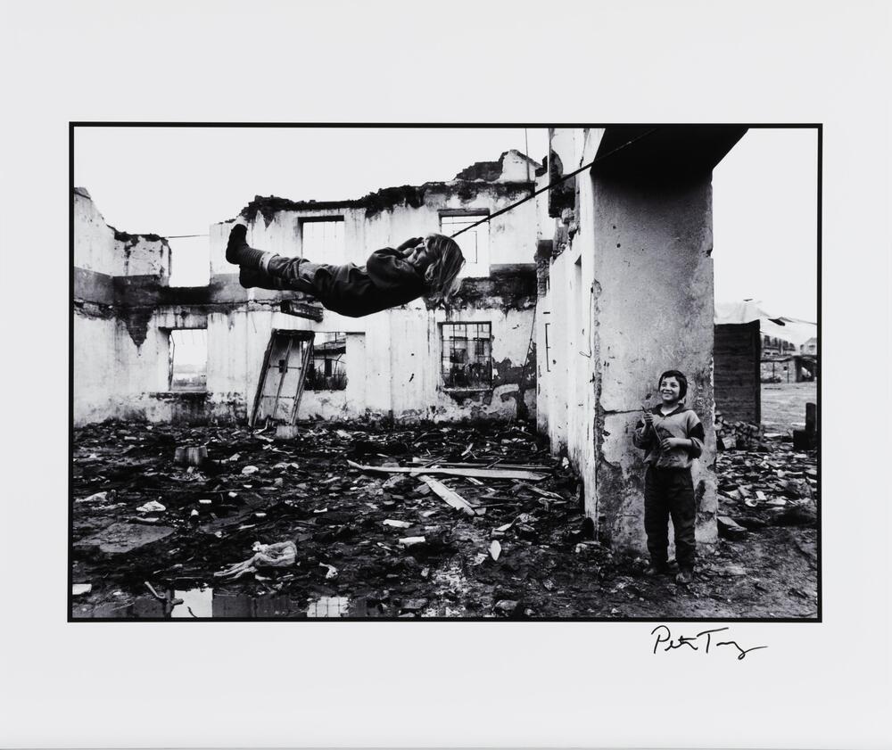 A black-and-white image of two children playing among a ruined, roofless structure and rubble. One child is swinging on a rope while the other watches.