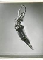 This photograph shows a ballerina leaping into the air with her arms above her head, in fifth position, toes pointed, and ankles crossed. 