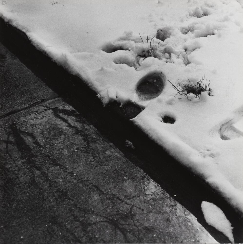 A black-and-white photograph of footprints in snow, appearing next to a puddle of water on pavement.