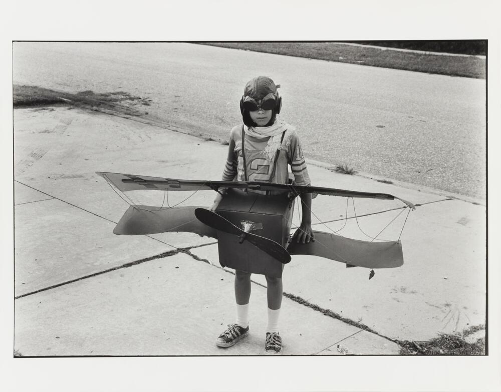 A black and white image of a boy wearing a scarf, aviation cap, and a cardboard boxed fashioned into an airplane. 