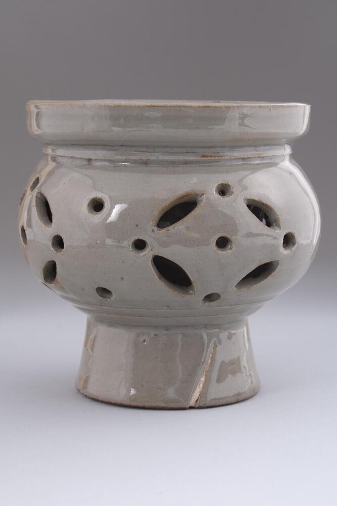 This white porcelain incense burner, featuring an openwork design on the body, is made of fine clay with high-iron content, which has tinged the surface with dark gray. The glaze has been removed from the part covered by the lid, exposing the red body. The foot is entirely glazed; sand spurs were supported in some parts of the foot rim during firing. Cracks formed in the foot and the base during firing. This object has a larger belly and narrower foot than typical traditional white porcelain incense burners.<br />
[Korean Collection, University of Michigan Museum of Art (2014) p.202]