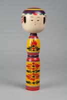 A wooden doll of three tiers made up of a head and body with no arms, legs or feet. Painted on the head is a face, hair, and headdress of yellow and different shades of red. The second and third tiers make up the body and have yellow, black, and red decorations.