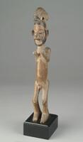 This carved, wooden Yaka figure depicts a man standing with an animal perched atop his head. The carving is stylized and exhibits characteristics typically seen among northern Yaka figural representations: flexed knees; arms bent with upturned palms positioned at shoulder level; and, an animal figure upon its head. In this case, the creature has a curved body and appears to be an anteater. The male figure has a narrow, cylindrical body; a slightly protruding belly; a simple coiffure; an elongated face; barely-open eyes from which vertical lines extend downward; a disproportionately large, pointed nose, and a darkened beard.