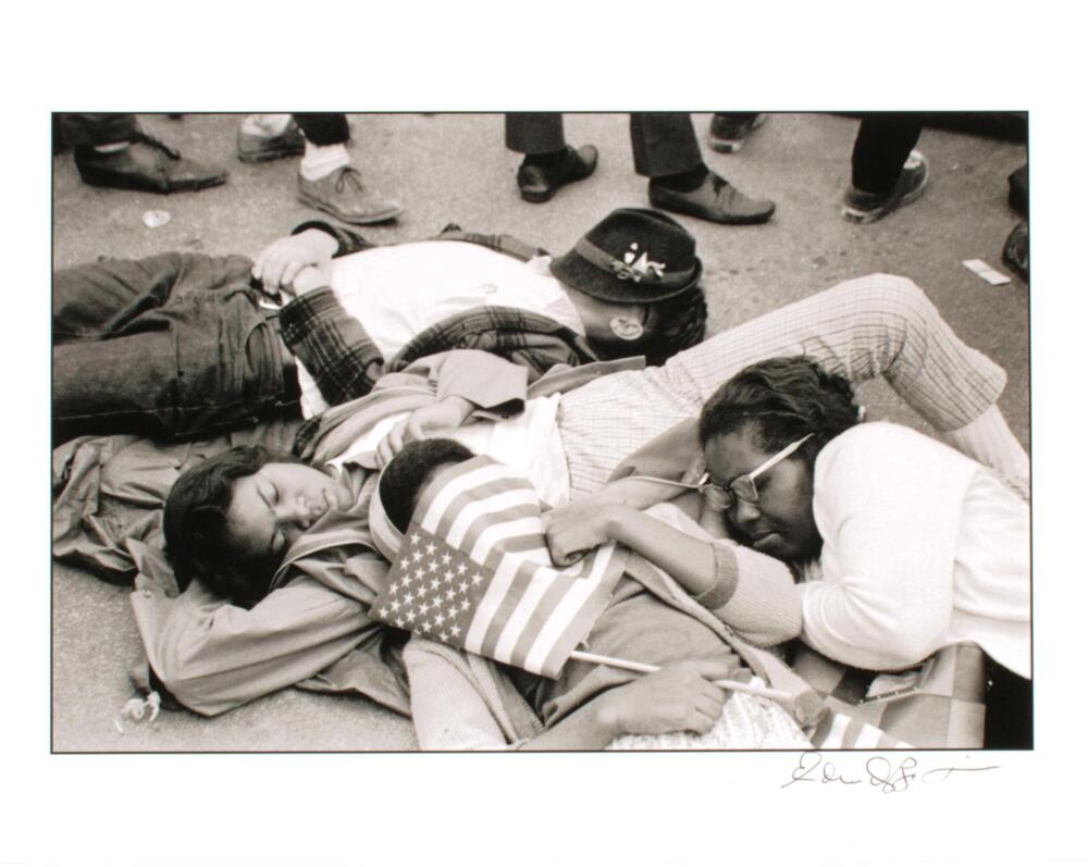 A black and white photograph of four people lying on the ground. One person's face is covered by an American flag, while the man behind her has his face covered by a hat. The group appears to be asleep. The feet and shoes of other people standing around the group of four is visible in the background. The print is signed (l.r.) "Edward Roberson" in pen.
