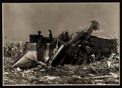 A photograph of a large farm machine in a field of corn. A man in a hat operates the machine.