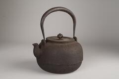 A round and squat kettle made of iron.  It has a tall, arching handle which is tapered at the ends of attachment and the handle on the cover is decorated with cut-outs.