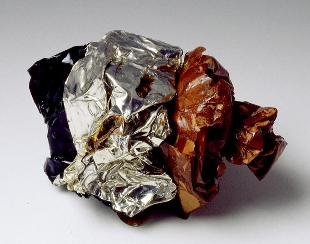 This small sculpture is made from crumpled paper and aluminum foil coated in resin. One end of the sculpture is blue, the other a brown-red, and the center is silver.  Pieces of paper which look like newsprint are stuck to the bottom of the sculpture. The piece is deceptively light.