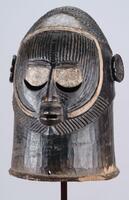 Dark colored mask with an ovoid shaped head. The face has incised parallel striations, round white eyes with open crescents below, round ears, a small nose and a small mouth. The coiffure is composed of parallel ridges circumventing the entire head. There are two holes around the bottom edge of the mask, one in the front and one in the back. 