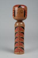 A wooden doll with two tiers made up of a head and body with no arms, legs or feet. Painted on the head is a face, hair, and red headdress. Painted on the body are red and black decorations.